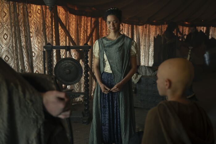 Young woman in medieval attire standing indoors with patterned curtains, related to Tanzyn Crawford's height compared to Tansell Too-Tall.