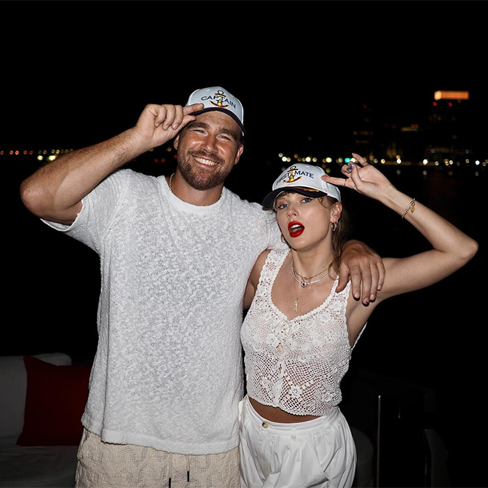 Taylor Swift and Travis Kelce wearing matching captain hats at night, posing closely with a city skyline in the background