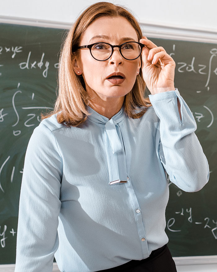 Female teacher in glasses looking surprised in a classroom, reacting to a mom struggling to contain her rage.