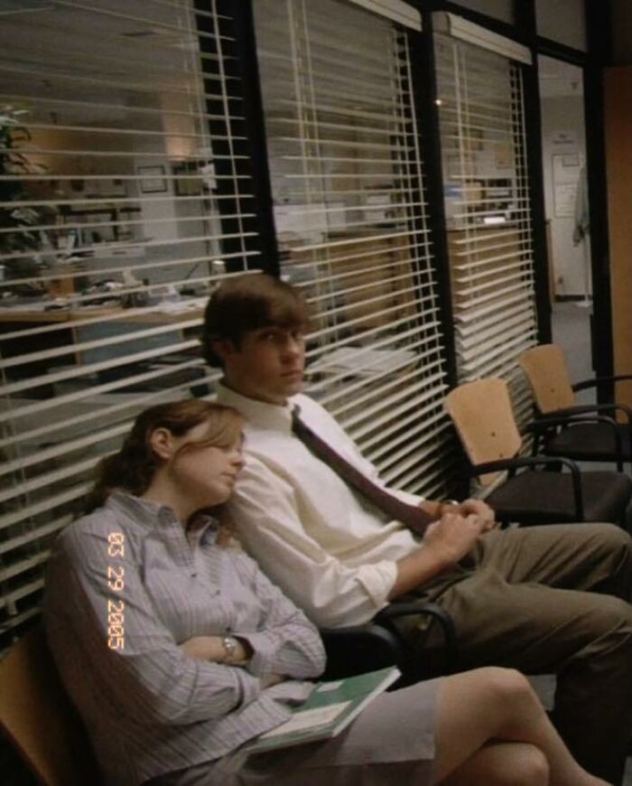 Two coworkers seated in an office waiting area, a woman resting her head on a man's shoulder in a meme inspired by The Office.
