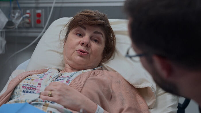 Woman lying in hospital bed, wearing patterned hospital gown, discussing with a man, relating to The Pitt Season 2.