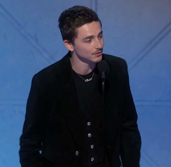 Timoth&eacute;e Chalamet speaking at a microphone during an event, linked to lip reader revealing Golden Globes fight.