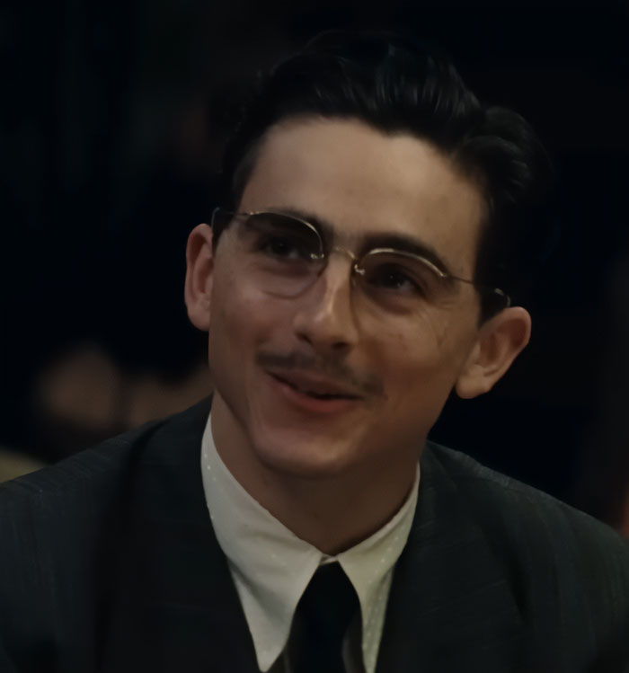 Young man wearing glasses and a vintage suit, smiling softly in a dimly lit setting related to Timoth&eacute;e Chalamet tone deaf message.