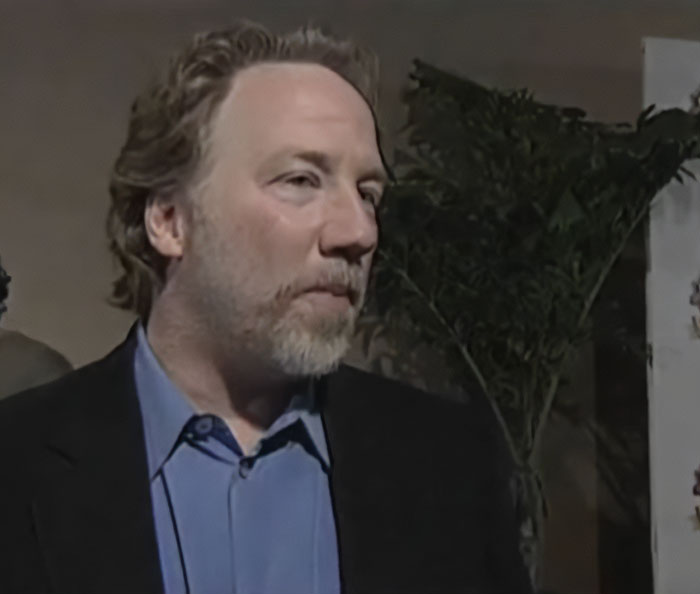 Man with beard and blue shirt standing indoors next to a plant, related to Melissa Gilbert and Timothy Busfield news.
