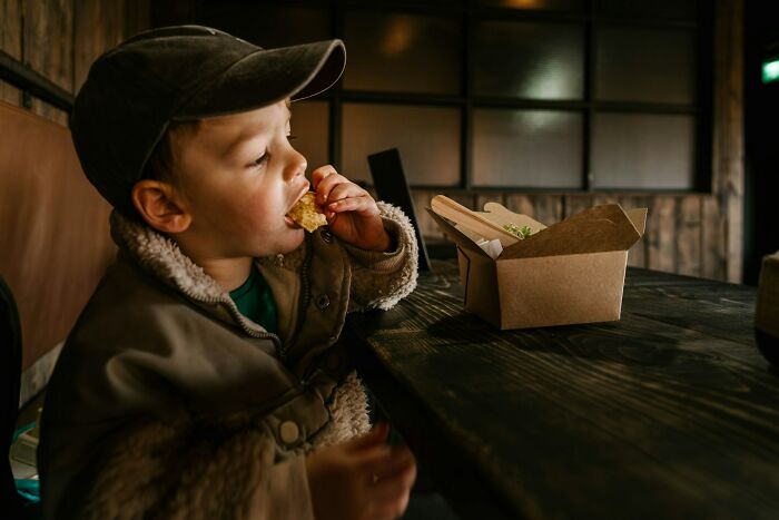 Young child wearing a cap eating food from a takeaway box at a wooden table in a casual service industry setting.