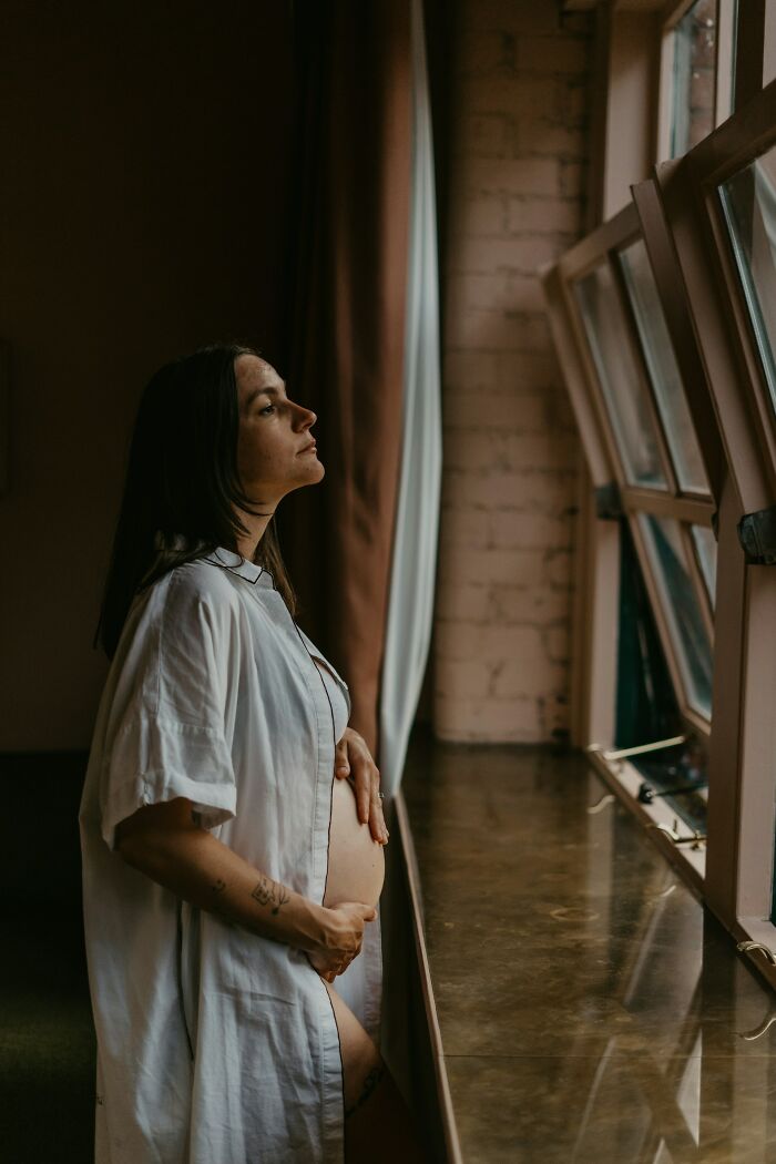 Pregnant woman in a white shirt looking out a window, reflecting on DNA test results and unexpected family discoveries.