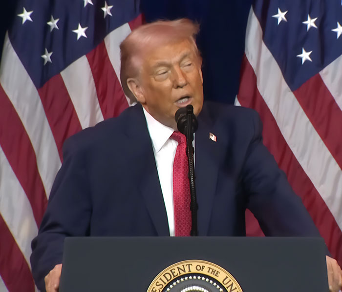 Donald Trump speaking at podium with American flags in background, showing pinkish hue in hair, professional stylist insight.