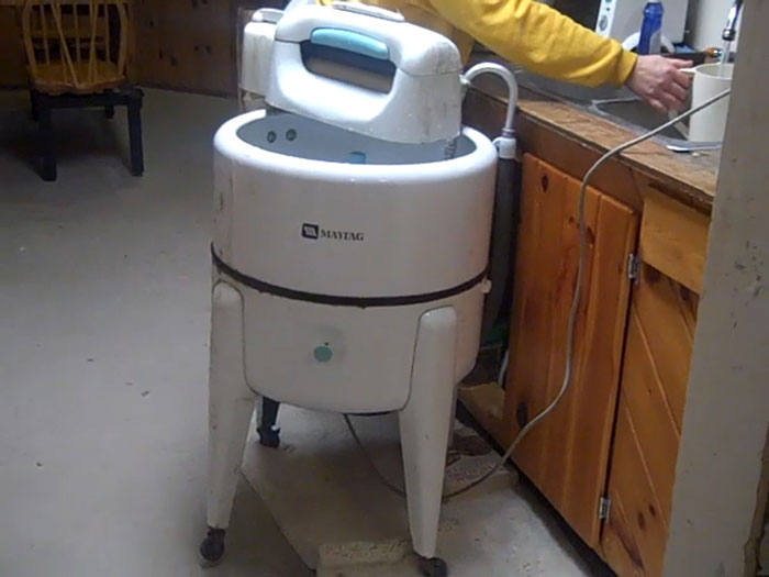 Vintage Maytag washing machine in a dim room with a person’s arm reaching over a wooden sink cabinet, unexplainable sight.