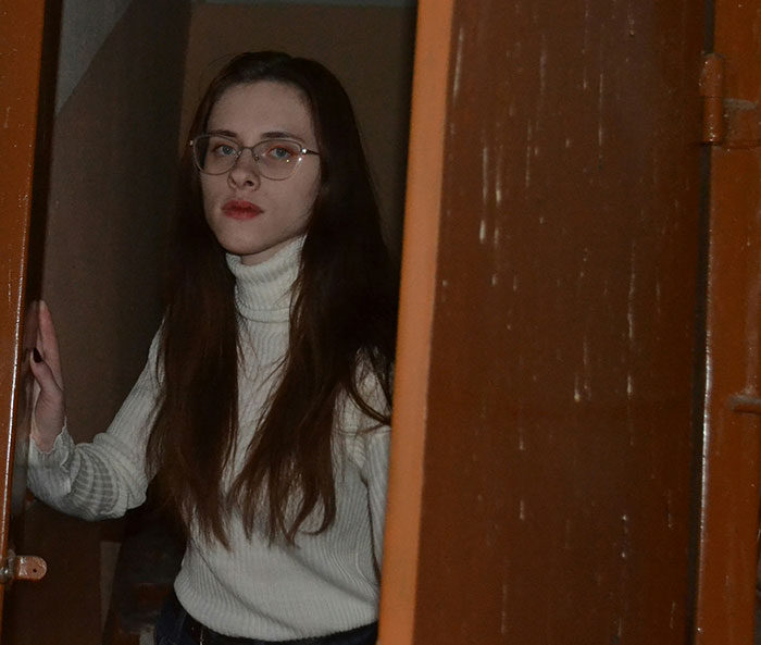 Young woman with glasses and long hair standing in a doorway, looking surprised by something unexplainable.