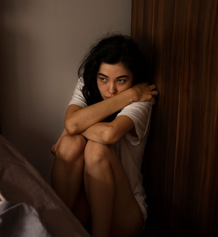 Young woman sitting in a dim room, hugging knees, looking thoughtful and unsettled, reflecting on unexplainable experiences.