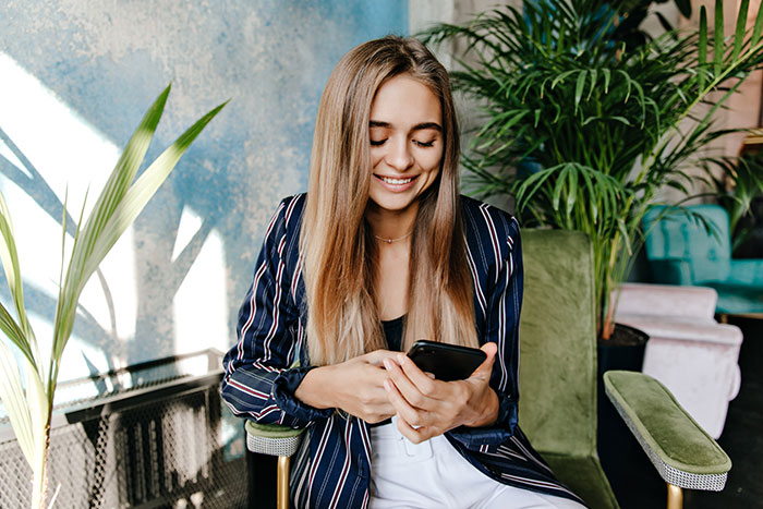 Young woman smiling while looking at phone indoors, illustrating guestzilla ignoring food allergy restrictions by party host.