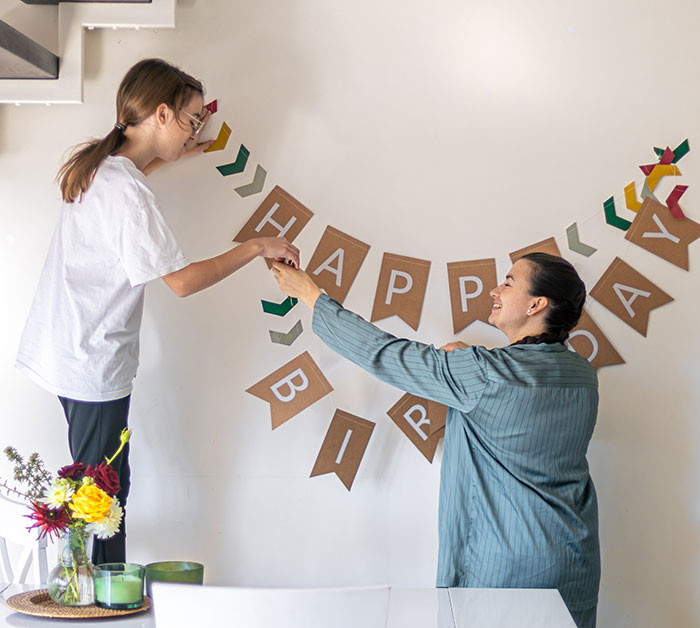 Two women decorating a birthday party banner, highlighting guestzilla ignoring food allergy restrictions set by host.