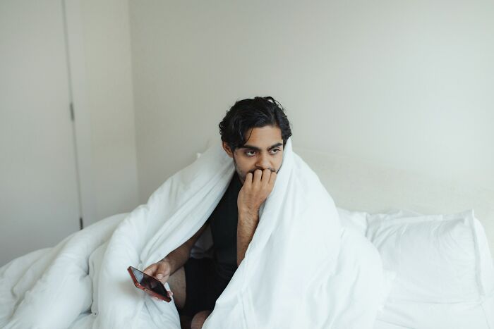 Young man wrapped in a blanket on bed, holding a phone, looking thoughtful, showing surprise or creeped out by intelligence.