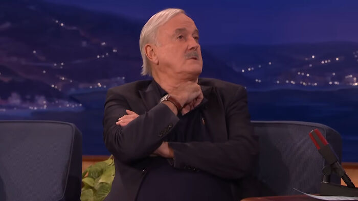 John Cleese sitting in a talk show set, engaged in conversation, illustrating moments people met their idols.