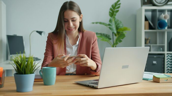Young woman in a pink blazer smiling at her phone at desk with laptop, illustrating worst coworkers at work stories.