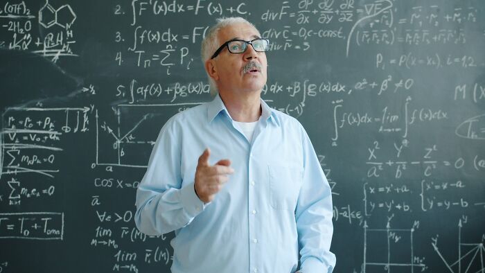 Older male teacher with glasses explaining math equations in front of blackboard showing weird teacher behavior in class.