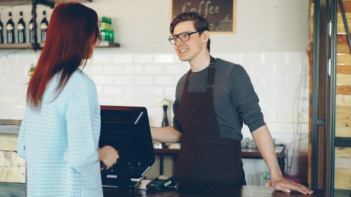 Barista smiling at customer across cafe counter, casual scene referencing It Rained Frogs