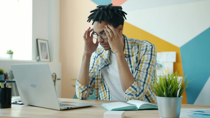 Young man wearing glasses, stressed while working on laptop, illustrating cultural differences in what’s normal and offensive.