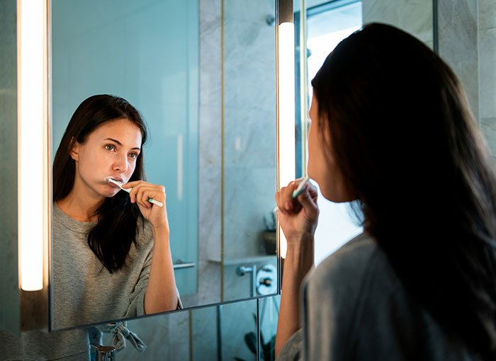 Woman brushing teeth in bathroom mirror, illustrating moments women share about attraction ending due to a weird ick.