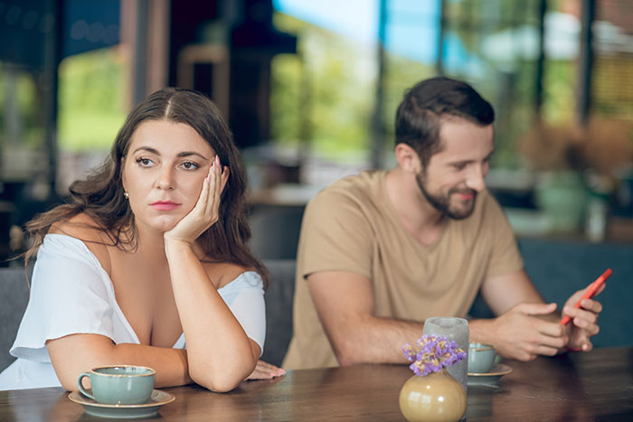 Woman showing ick feeling while sitting at a table with man who is distracted by his phone in a cafe setting.