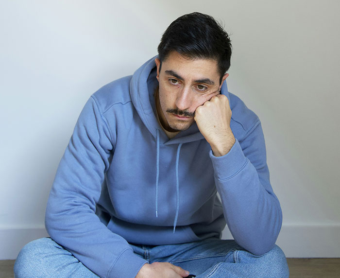 Man in blue hoodie sitting on floor, looking devastated and deep in thought after discovering his wife's infidelity.