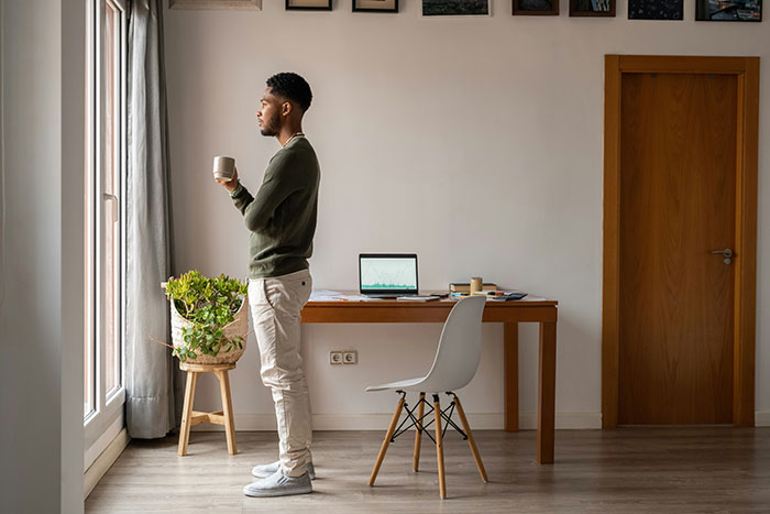 Man standing by window with coffee, looking devastated after snooping on wife's conversation about infidelity.