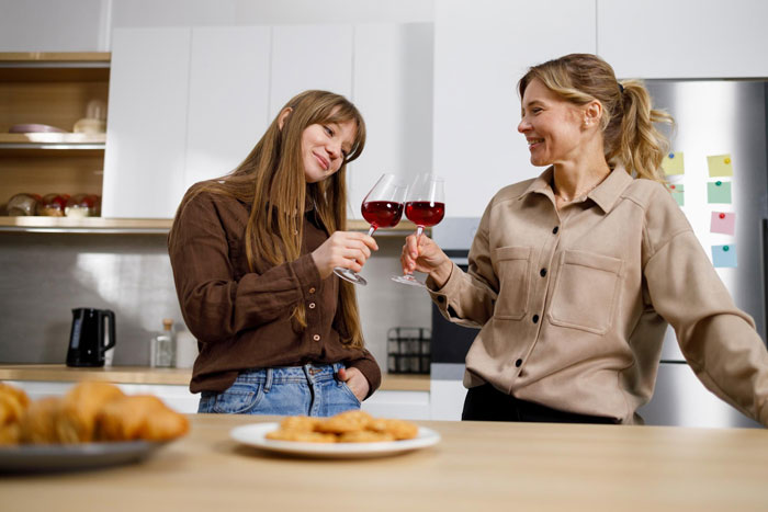 Two women in a kitchen toasting red wine over pastries, smiling and chatting &mdash; portraying a lesbian bestie moment