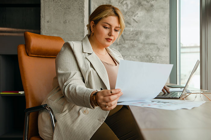 Young mom reviewing documents at desk, appearing thoughtful and focused while working on her laptop near a window.