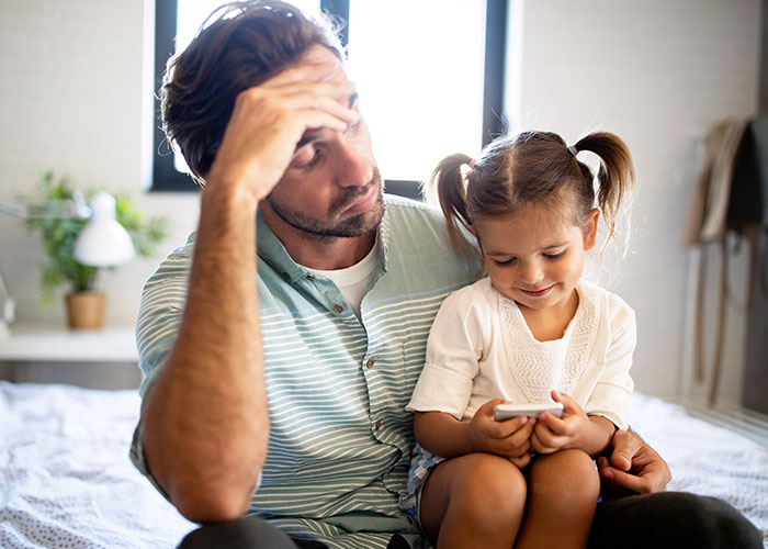Man looking stressed while sitting with young daughter playing on phone, illustrating unpaid nanny feelings after partner leaves child.