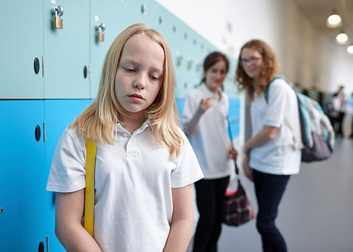 Sad girl standing by school lockers while two girls in the background appear to be bullying her in a hallway.