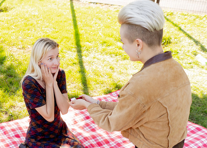 A person proposing outdoors on a red plaid blanket with a surprised fianc&eacute;e showing an emotional reaction.