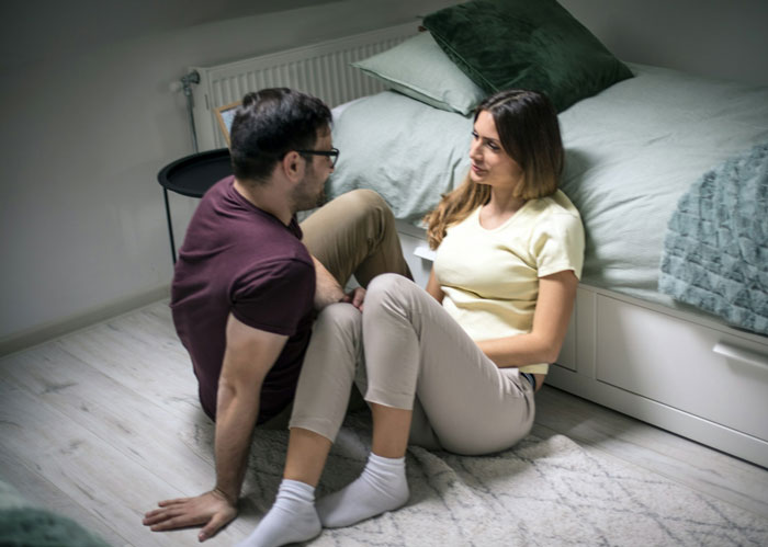 Mom of 6 stressed and overwhelmed sitting on floor talking to man in a bedroom with a bed in the background