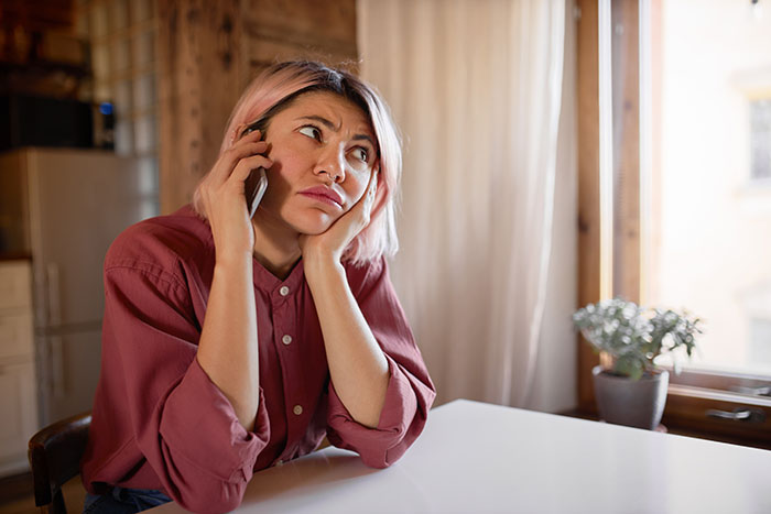 Frustrated woman on phone at kitchen table, pink shirt, upset expression, nearby plant, mention of heated blanket gift Frustrated woman on phone at kitchen table, pink shirt, upset expression, nearby plant, mention of heated blanket gift