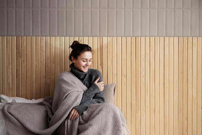 Young woman smiling wrapped in a heated blanket on a bed, cozy grey throw against wood-paneled wall Young woman smiling wrapped in a heated blanket on a bed, cozy grey throw against wood-paneled wall