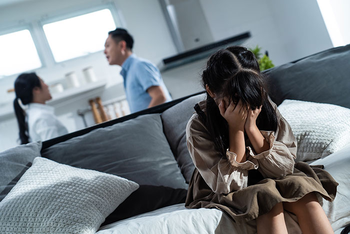 Woman covering face in distress on couch while parents argue in background, illustrating conflict over kids and trip to Italy. Woman covering face in distress on couch while parents argue in background, illustrating conflict over kids and trip to Italy.