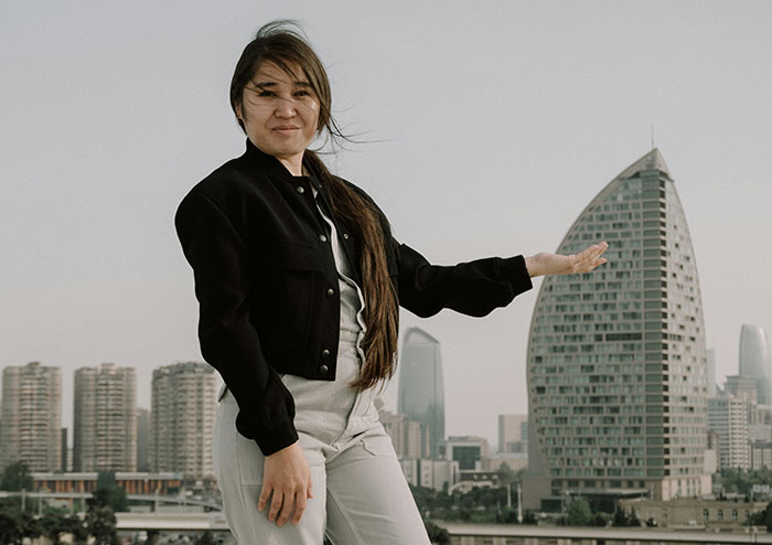 Woman standing outdoors in casual clothes, gesturing towards a city skyline in a modern urban setting, discussing obsessions.