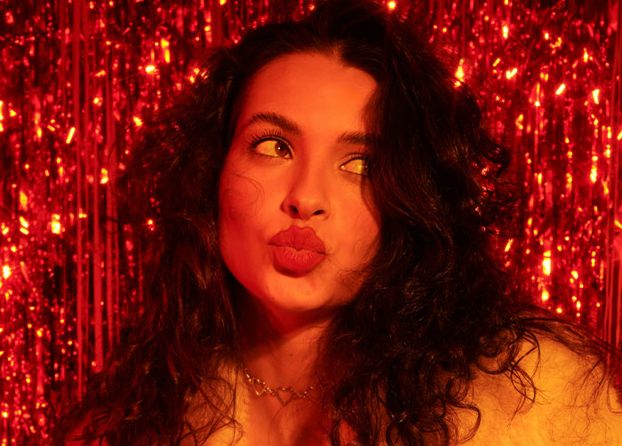 Young woman with curly hair posing playfully against a red shimmering background about what women obsess over.