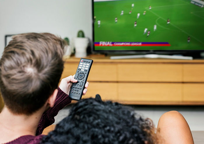 A man holding a remote control watching a soccer match on TV, highlighting what women obsess over for no reason.