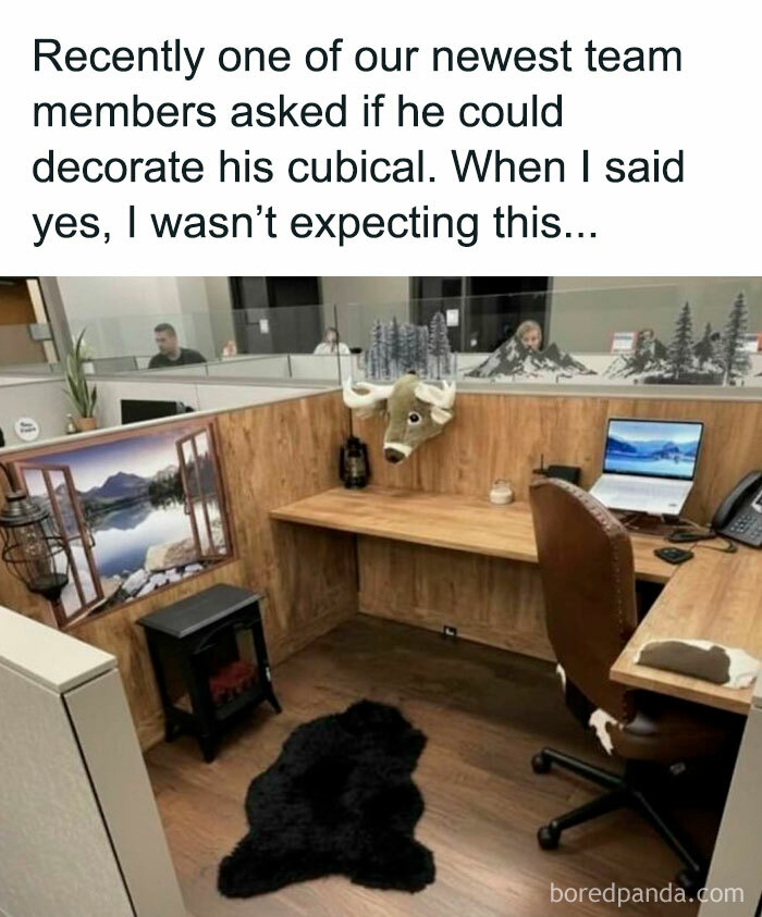 Office cubical decorated with a mounted animal head, faux fireplace, black rug, and scenic outdoor wall decal work memes.