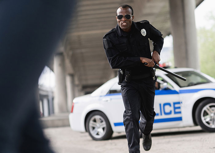 Police officer wearing sunglasses, holding a baton, running near a police car in the wrong place at the wrong time scenario.