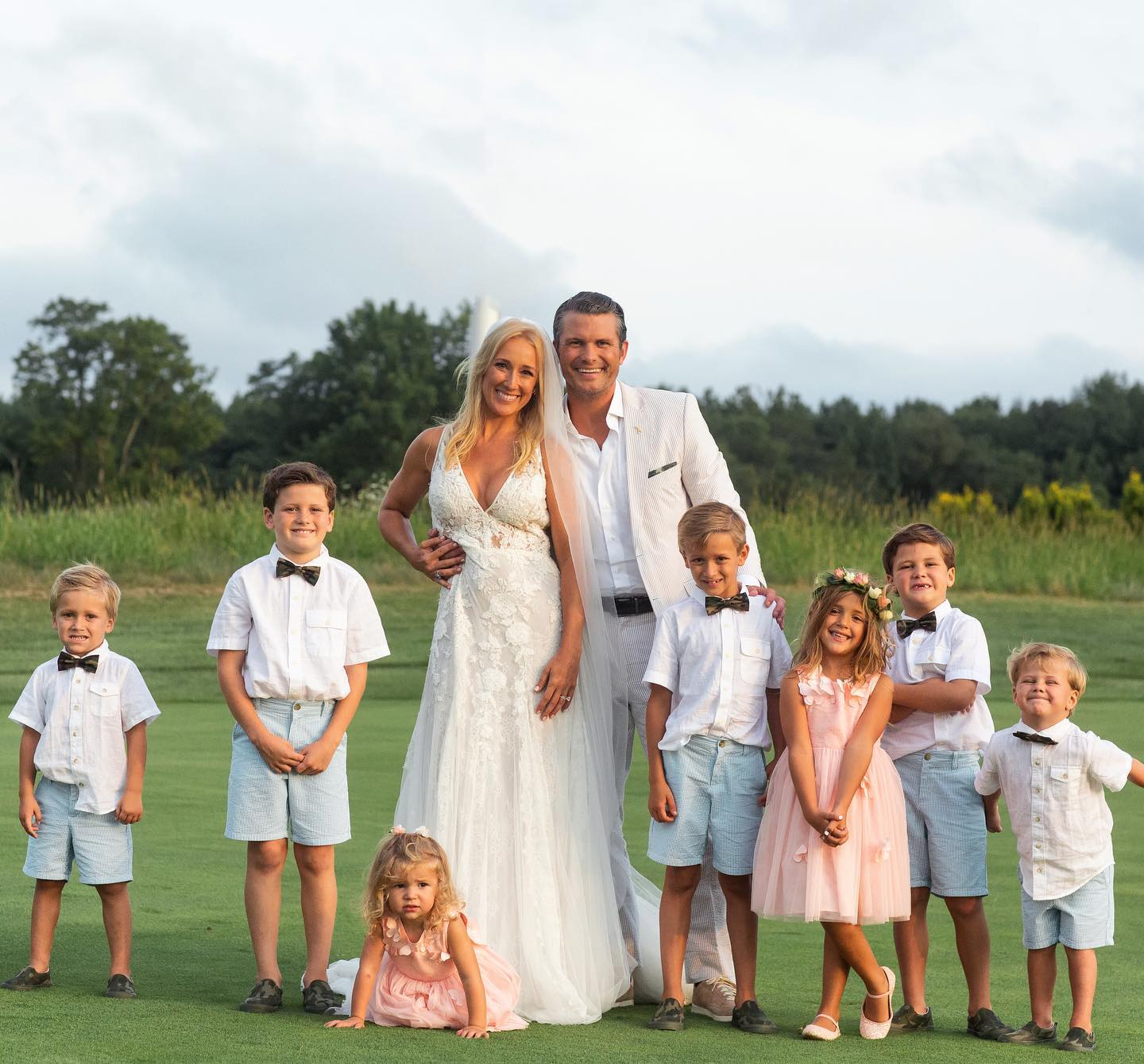 Pete Hegseth and family dressed in formal attire outdoors, smiling with seven children on a green lawn.