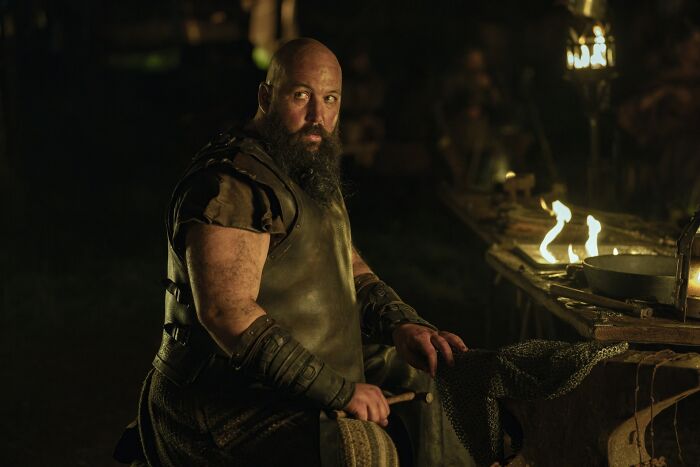 A bearded man in medieval armor sitting by a firelit table, referencing Tanzyn Crawford height and Tansell Too-Tall.