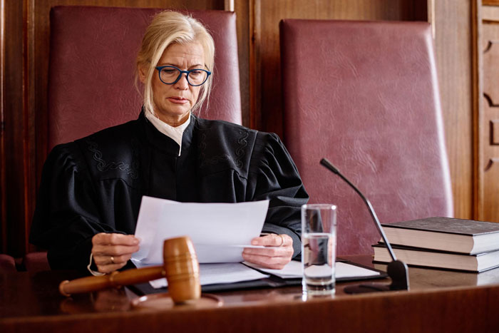 Female judge wearing glasses and reading legal documents at courtroom desk with gavel, books, and microphone.