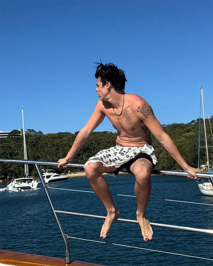 Yungblud shirtless on yacht railing, showing tattoos and wearing shorts during a sunny day near water and boats.