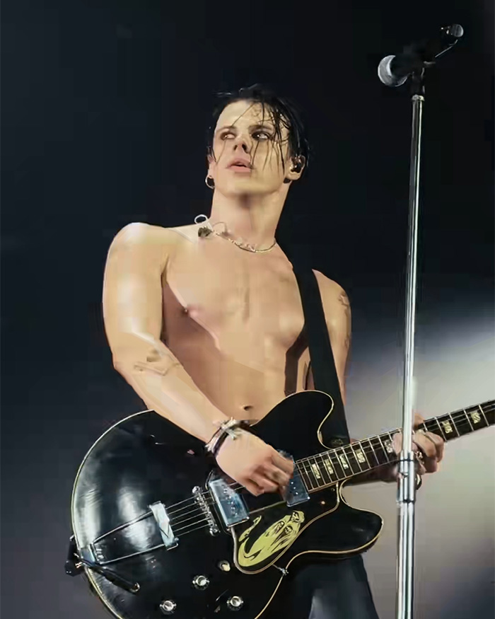 Yungblud shirtless playing electric guitar on stage with microphone nearby, spotlight highlighting his performance.