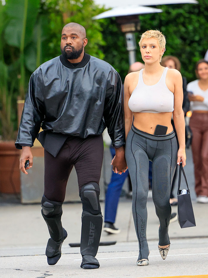 Bianca Censori sai com top de lingerie e legging enquanto dá a mão a um homem de roupa preta ao ar livre.