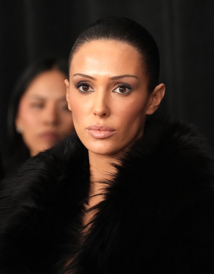Bianca Censori com penteado elegante e maquiagem ousada, vestindo casaco de pele preto em ambiente de evento de moda.
