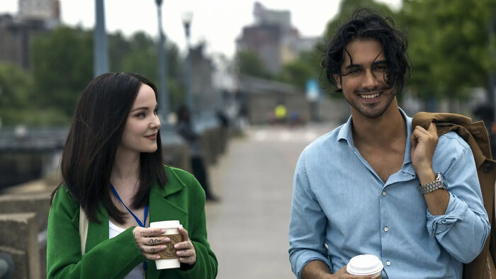 Two characters from 56 Days smiling and walking outdoors, each holding a coffee cup in a casual setting.