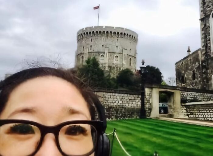 Woman wearing glasses and headphones taking a chaotic selfie in front of a historic castle capturing mom energy on social media.