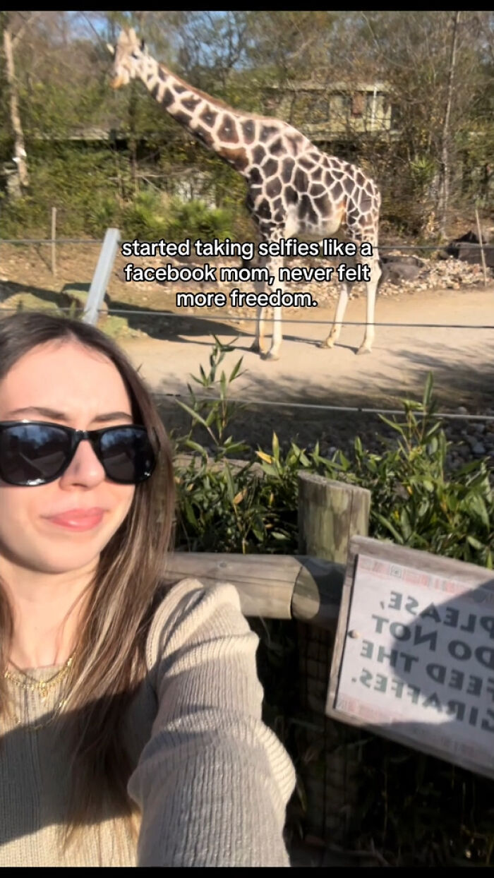 Young woman wearing sunglasses taking a hilarious mom selfie at a zoo with a giraffe and social media chaotic energy.